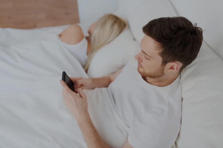 trouble erection Homme brun allongé dans sont lit qui regarde son téléphone portable à côté d'une femme blonde qui dort
